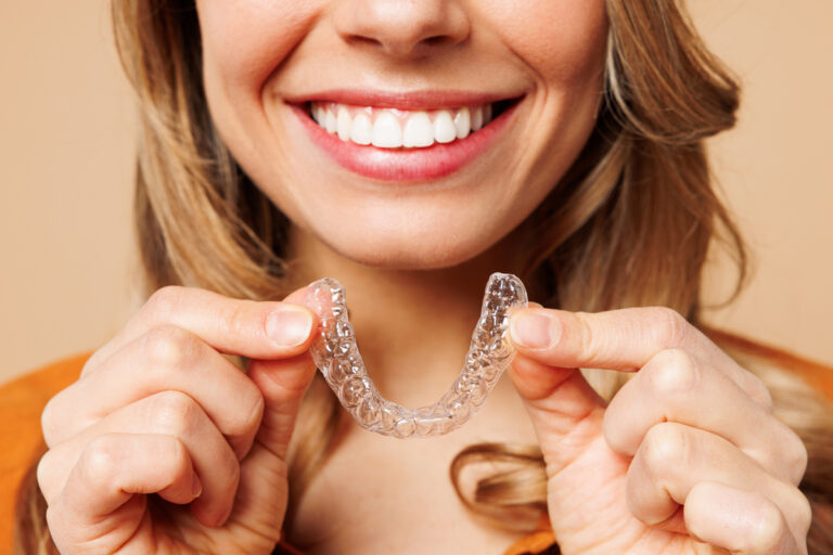 Close up cropped young woman wear orange shirt casual clothes hold in hand invisible transparent aligners, invisalign bracer smile isolated on plain pastel light beige background. Lifestyle concept.