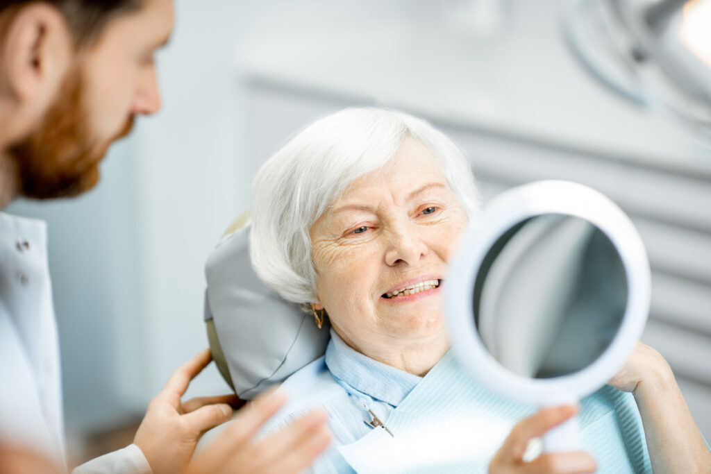 Happy elderly woman enjoying her beautiful toothy smile looking to the mirror in the dental office